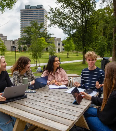 Vijf studenten zitten aan een picknicktafel op de campus. In de achtergrond is het Erasmusgebouw te zien.
