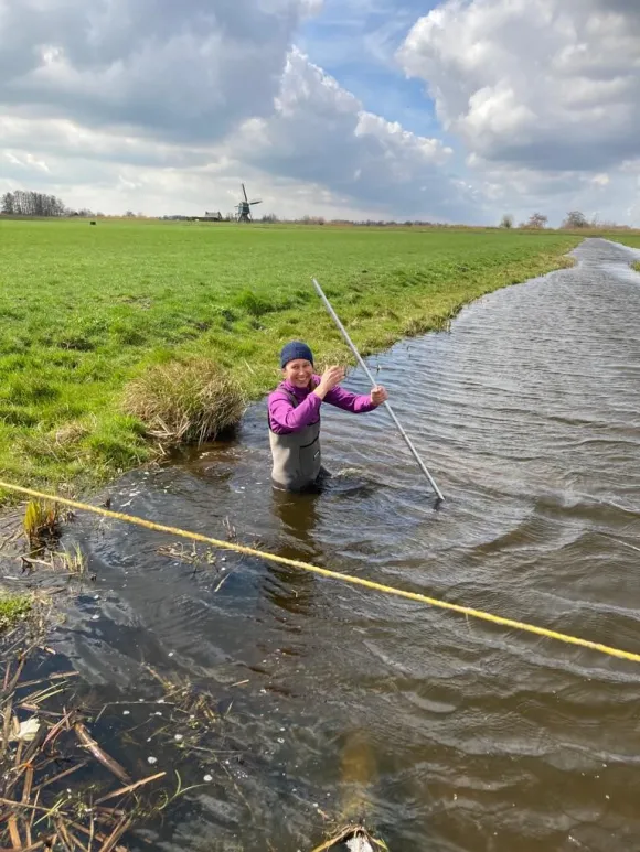 Sarian Kosten standing in a ditch next to a green field of grass, with a Dutch windmill in the background
