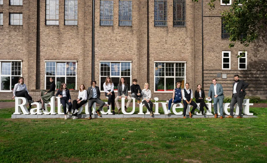 Een groepsfoto van de Universitaire Studentenraad van 2023-24. Ze zitten voor het Berchmanianum op de witte letters die samen Radboud Universiteit.