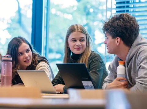 Foto van drie studenten in een klaslokaal met twee laptops voor zich.