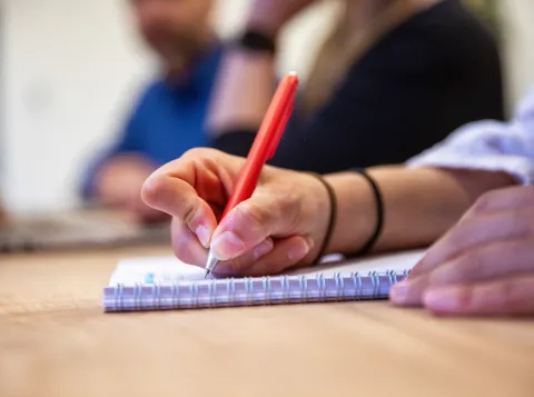 Een ingezoomde foto van een hand die een rode pen vasthoudt en op een kladblok schrijft.