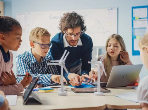 Basisschool klaslokaal: enthousiaste leraar met tabletcomputer legt aan briljante jonge kinderen uit hoe windturbines werken.