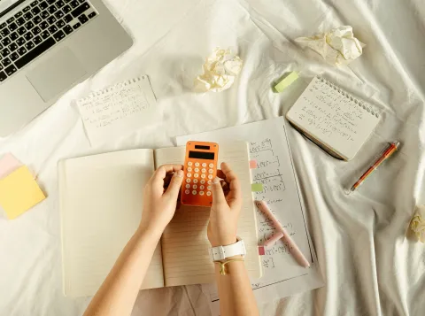 Hands holding a calculator, with a math notebook open and a laptop on the side. 