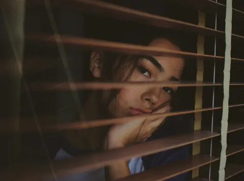 Portret van somberkijkende vrouw achter jaloezieën