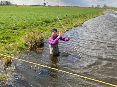 Sarian Kosten standing in a ditch next to a green field of grass, with a Dutch windmill in the background