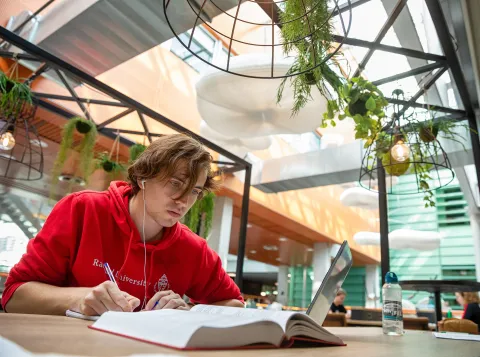 Een student zit te studeren in het Huygensgebouw met een open boek voor zich. Hij draagt een rode Radboudhoodie, heeft oortjes in en houdt een pen vast.