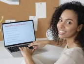 Vrouw kijkt in de camera terwijl ze achter een laptop zit
