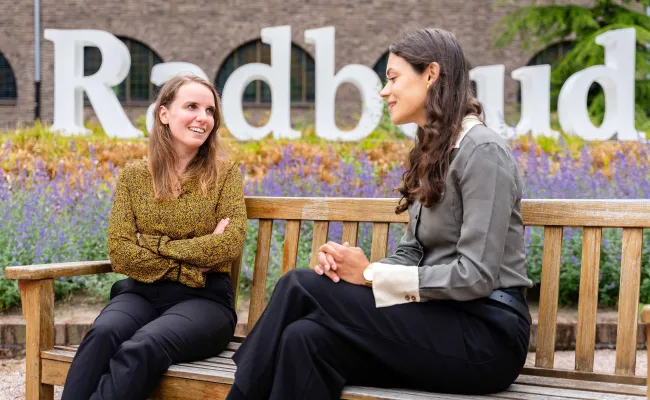 Twee vrouwen zitten op een bankje te overleggen met de 'Radboud' letters in de achtergrond.