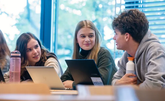Foto van drie studenten in een klaslokaal met twee laptops voor zich.
