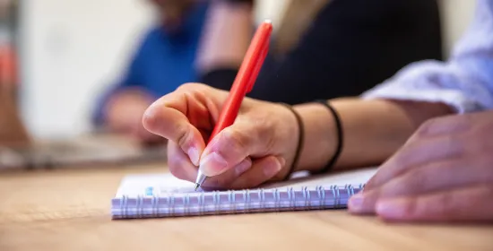 Een ingezoomde foto van een hand die een rode pen vasthoudt en op een kladblok schrijft.