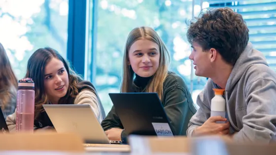 Foto van drie studenten in een klaslokaal met twee laptops voor zich.