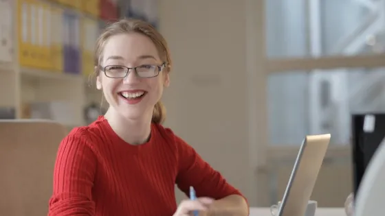 Vrouw achter laptop die lachend de camera inkijkt