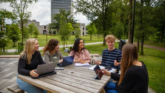 Vijf studenten zitten aan een picknicktafel op de campus. In de achtergrond is het Erasmusgebouw te zien.